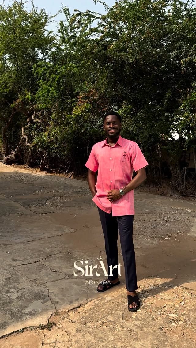 Man standing outdoors in a salmon-pink Adinkra shirt and dark trousers